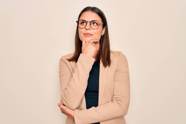 jeune belle femme d’affaires brune utilisant la veste et les glaces au-dessus du fond blanc avec la main sur le menton pensant à la question, expression pensive. sourire avec le visage pensif. concept de doute. - contemplation photos et images de collection