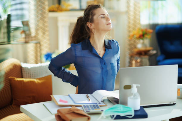 tired woman in modern house in sunny day having back pain tired modern woman in blue blouse in the modern house in sunny day having back pain in temporary home office during the coronavirus epidemic. backache photos stock pictures, royalty-free photos & images