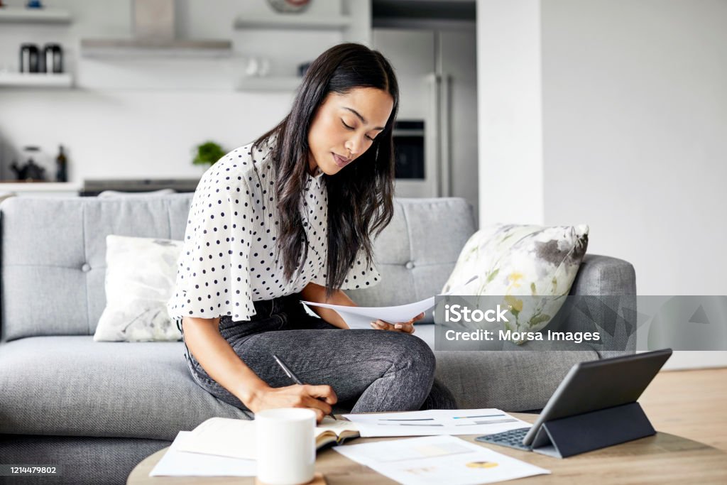 Woman analyzing documents while sitting at home Young woman analyzing bills while writing in diary. Beautiful female is using digital tablet at table. She is sitting on sofa at home. Working At Home Stock Photo