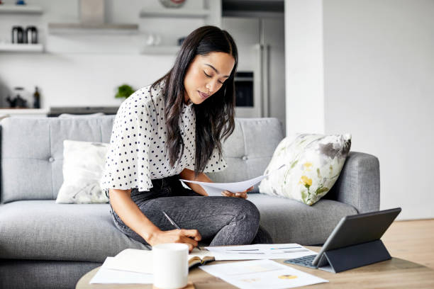 Woman analyzing documents while sitting at home Young woman analyzing bills while writing in diary. Beautiful female is using digital tablet at table. She is sitting on sofa at home. financial bill stock pictures, royalty-free photos & images