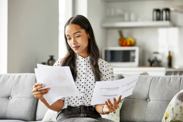 Woman analyzing financial documents at home Young woman checking financial documents. Female is analyzing bills in living room. She is sitting on sofa at home. paperwork stock pictures, royalty-free photos & images