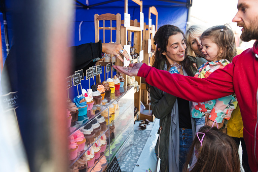 Side view of a woman holding her daughter while the daughter's father is paying for a cupcake she has just chose from a stall at the Whitley Bay Food Festival.