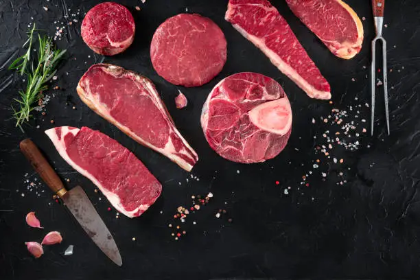 Photo of Various cuts of meat, shot from the top on a black background with salt, pepper, rosemary and knives, with copy space
