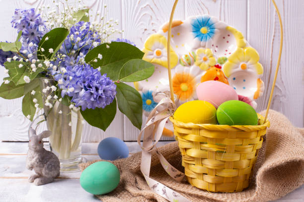 composizione pasquale con uova colorate in un cesto di vimini, una statuina di coniglio e un bouquet primaverile in un vaso e un bellissimo piatto di ceramica su un tavolo di legno bianco - easter egg figurine easter holiday foto e immagini stock