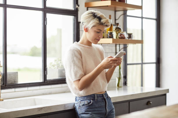 A woman using smart phone in kitchen Smiling A woman using smart phone. Female is text messaging while leaning on countertop. She is at home. shorthair stock pictures, royalty-free photos & images