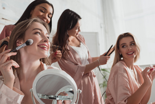 Selective focus of multiethnic women putting makeup with cosmetic brush and doing hairstyles in room at bachelorette party