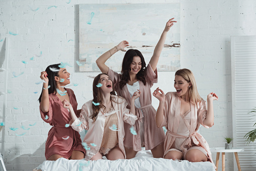 Happy and excited multiethnic women with hands in air on bed with feathers in room at bachelorette party