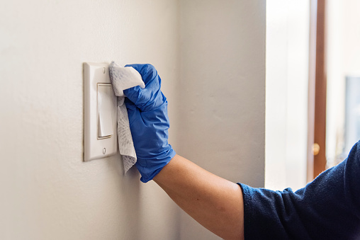 Female hand with blue glove wiping light switch with disinfectant wipe. Horizontal indoors close-up with copy space.