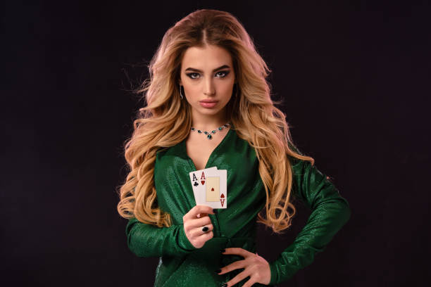 blondynka modelka w zielonej stylowej sukience i biżuterii. połóż rękę na biodrze, pokazując dwie karty do gry, pozując na czarnym tle. poker, kasyno. zbliżenie - casino black and white gambling chip gambling zdjęcia i obrazy z banku zdjęć