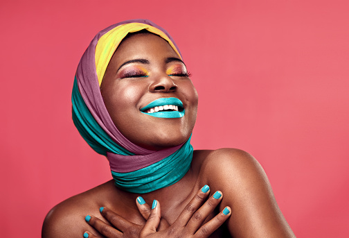 Studio shot of a beautiful young woman smiling while wearing a head wrap and make up against a pink background