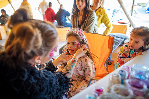 A young girl getting glitter painted on her face by a face painter. She is sitting at a stall, part of the Whitley bay Food Festival while her family watch.