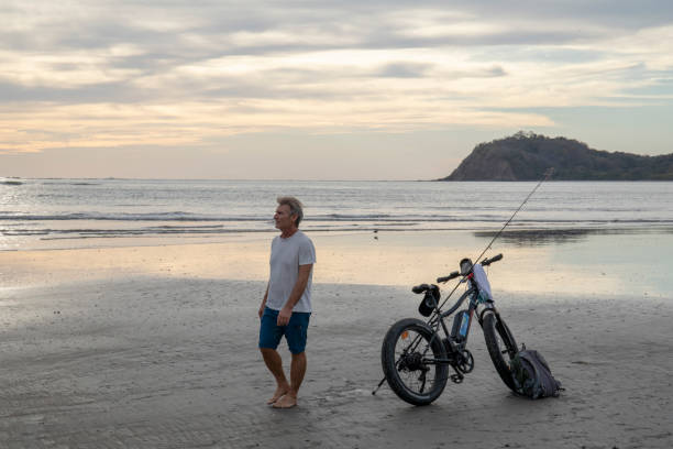 homem com e-bike caminha pela praia tropical - 16723 - fotografias e filmes do acervo