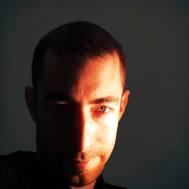 istock Bearded young man with a red light on his half-face looking at the camera with a malicious smile in the darkness 1214532245