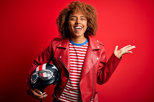 Young African American motocyclist woman holding motorcycle helmet over red background very happy and excited, winner expression celebrating victory screaming with big smile and raised hands