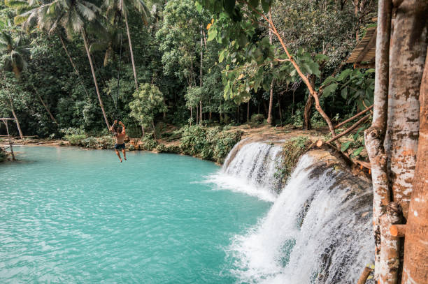A young caucasian tattooed man plays with a rope swing over the Cambugahay Falls in Siquijor Island, Philippines A man playing with a rope swing in Cambugahay Falls siquijor island stock pictures, royalty-free photos & images
