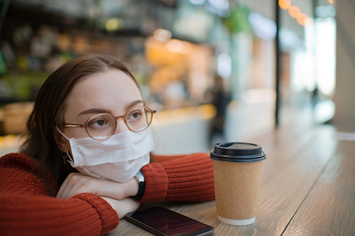 Teenager wearing medical mask protecting herself against virus in a food court of a shopping mall or airport lobby with reusable coffee cup