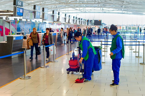 Cleaning women in uniform with broom, scoop and buckets in the international airport terminal, row of people at passport control in background