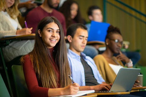A group of multi-ethnic students sit in a lecture hall and take notes and focus during class.