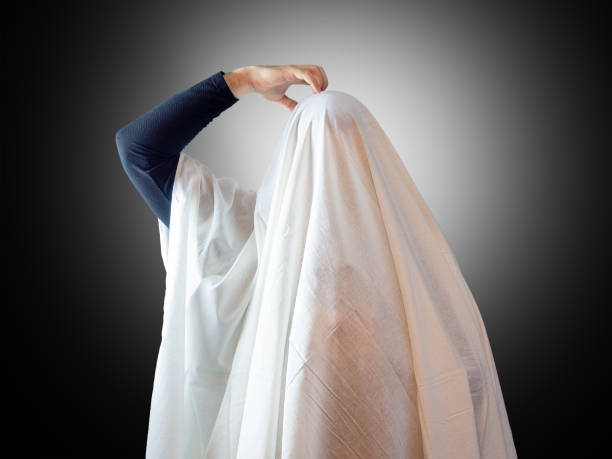 istock A man, covered with a white sheet like a ghost, scratches his head with his hand on a dark background with backlight 1214546628
