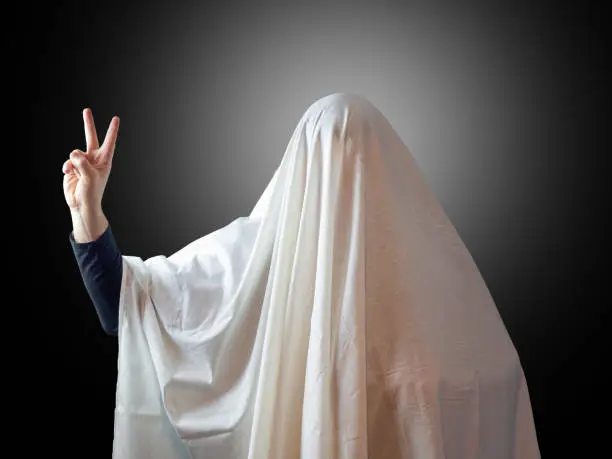 Photo of A man covered with a sheet like a ghost shows two fingers indicating victory on a dark background with backlight in the background