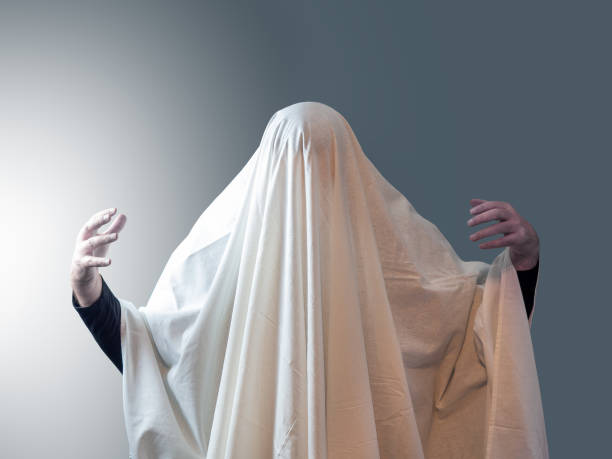 istock The man, covered in white drapery like a ghost, pulled his hands out from under it against a gray background with light on the left 1214546635