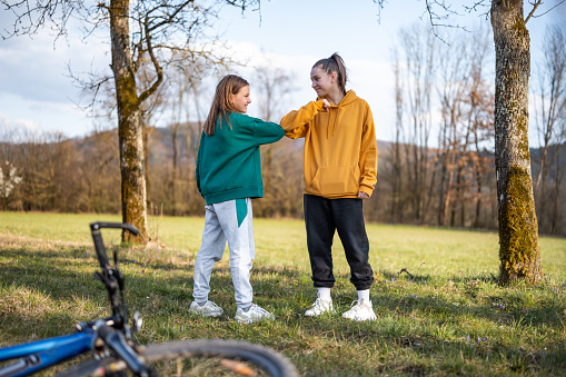 Two caucasian school girls in sport clothes using elbow bump as an alternative handshake outdoors in nature. There is a bicycle on the ground.
