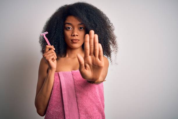 진지하고 자신감있는 표정으로 정지 표지판을하고 있는 열린 손으로 여성 면도기의 여성 면도칼을 들고 수건에 아프로 머리를 가진 젊은 아프리카 계 미국인 여성, 방어 제스처 - razor 뉴스 사진 이미지