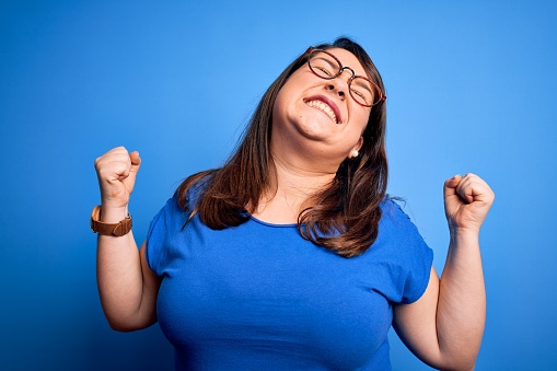 Beautiful brunette plus size woman wearing casual t-shirt over isolated blue background very happy and excited doing winner gesture with arms raised, smiling and screaming for success. Celebration concept.