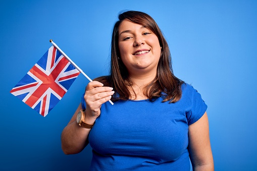 Beautiful plus size woman holding united kingdom flag over blue background with a happy face standing and smiling with a confident smile showing teeth