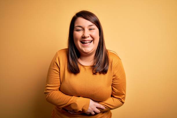 Beautiful brunette plus size woman wearing casual sweater over isolated yellow background smiling and laughing hard out loud because funny crazy joke with hands on body. Beautiful brunette plus size woman wearing casual sweater over isolated yellow background smiling and laughing hard out loud because funny crazy joke with hands on body. people laughing hard stock pictures, royalty-free photos & images