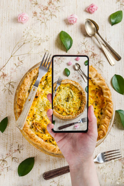Person taking photos with his mobile to a delicious quiche surrounded by flowers, leaves and rustic forks and spoons in a fresh, spring setting stock photo