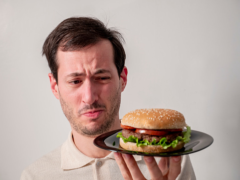Handsome young man with disgusted facial expression, holding tasty hamburger