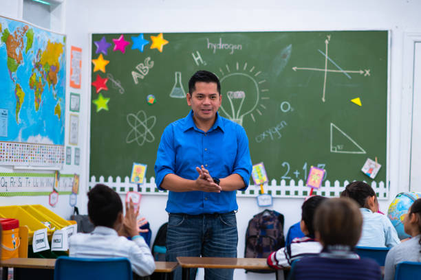A mixed-race male teacher stands in front of his class of multi-ethnic students as they sit with their backs to the camera and focus on his instruction. The teacher is standing in front of the blackboard and map on the wall.