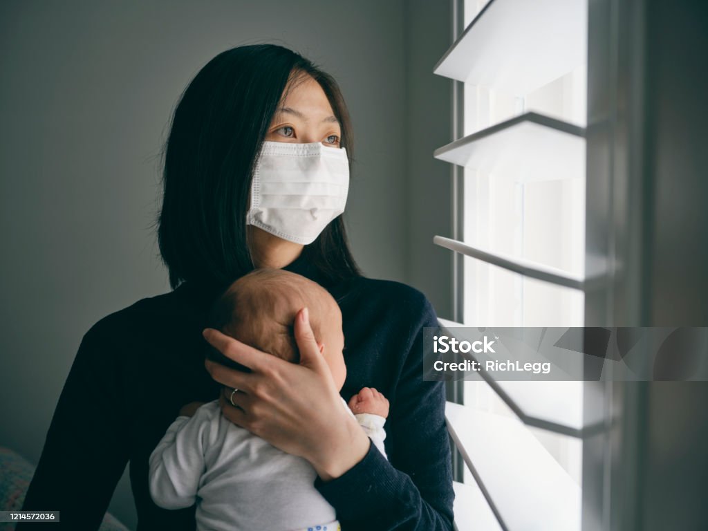 A mother and baby in home isolation from infectious disease. A mother wearing a surgical mask with her baby in home isolation from infectious disease. Baby - Human Age Stock Photo
