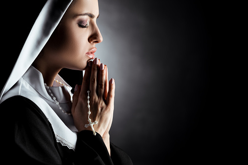 beautiful young nun with closed eyes praying with cross on grey