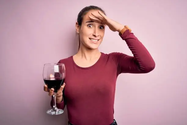 Photo of Young beautiful woman drinking glass with red wine over isolated pink background stressed with hand on head, shocked with shame and surprise face, angry and frustrated. Fear and upset for mistake.