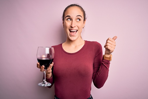 Young beautiful woman drinking glass with red wine over isolated pink background pointing and showing with thumb up to the side with happy face smiling
