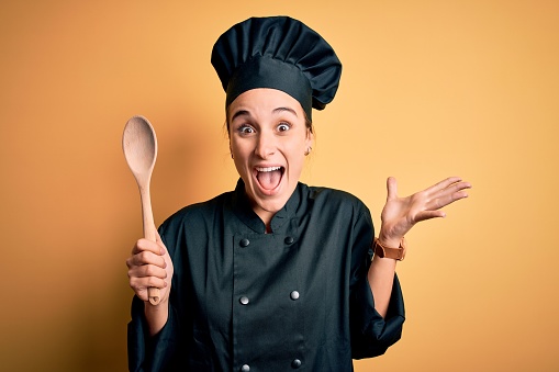 Young beautiful chef woman wearing cooker uniform and hat holding wooden spoon very happy and excited, winner expression celebrating victory screaming with big smile and raised hands