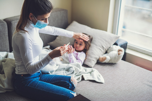 Mother wearing protective mask at home and measure a temperature on her sick child.