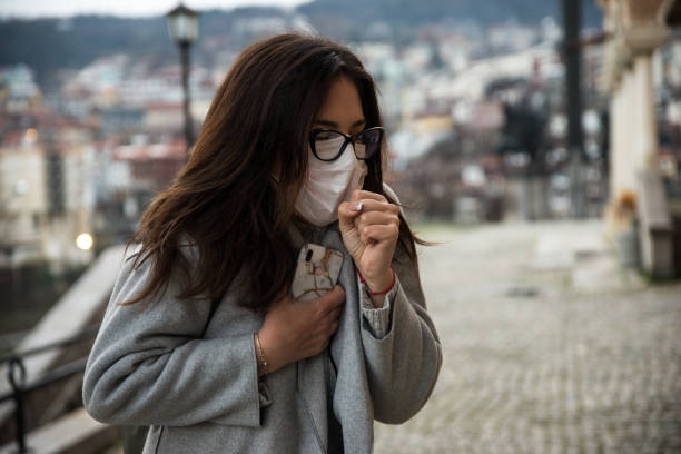 jovem mulher usa máscara facial e tosse enquanto está de pé na cidade - smog city pollution town - fotografias e filmes do acervo