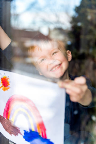 Young Boy sticking his drawing on home window during the Coronavirus Covid-19 crisis, Many people are putting a rainbow to tell neighbors that people inside this house are ok. #Stay at home.