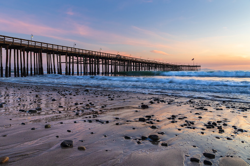 Ventura Pier at sunset, Ventura, California. Rocks and sand in foreground, water receded in low tide, with incoming waves. Lamps on pier; colored twilight sky in background.