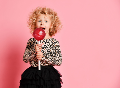 Pretty happy curly blonde hair young little child girl hold huge sweet lollypop candy surprised screaming yelling on pastel color pink background