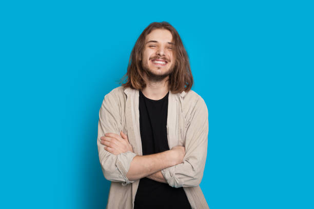 uomo caucasico fiducioso in posa con le braccia incrociate mentre sorride su uno sfondo blu - capelli lunghi foto e immagini stock