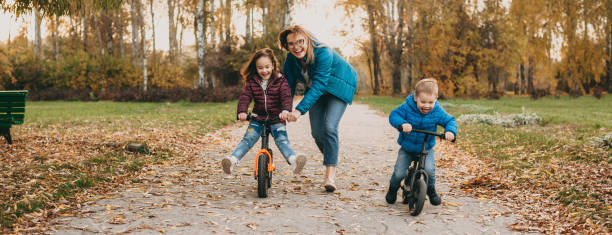 smiling caucasian mother is playing with her small kids while teaching them to ride the bike - glasses child cute offspring imagens e fotografias de stock