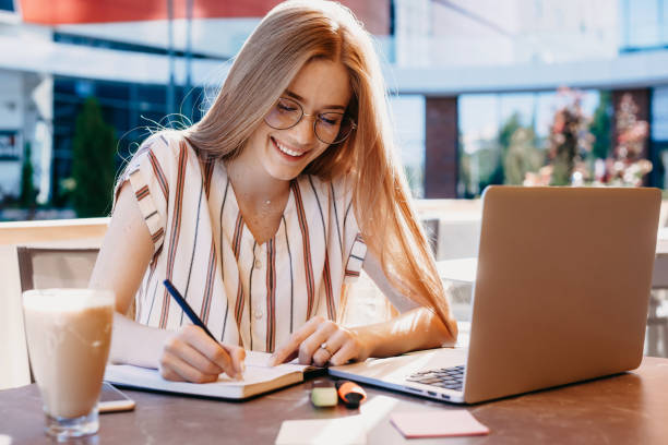 istock Ginger caucasian student with freckles is writing something in her notebook while drinking a coffee and using her laptop 1214624200