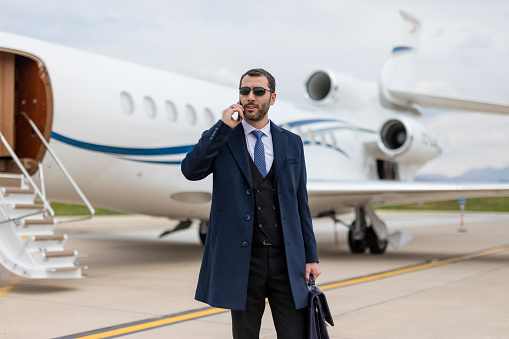 Man wearing elegant clothes walking away from private jet airplane and talking over mobile phone.
