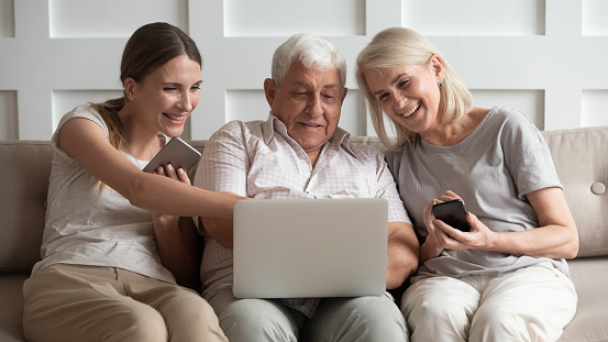 Happy older parents with adult daughter using electronic devices, phones, looking at laptop screen, smiling grandparents and granddaughter watching funny video or reading news in social network