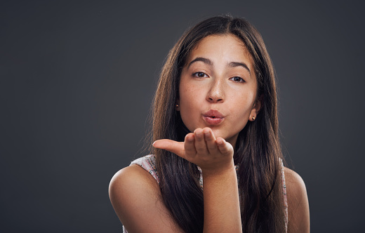 Cropped portrait of an attractive teenage girl standing alone and blowing kisses against a dark studio background