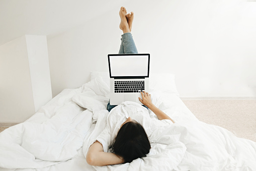 Happy stylish girl lying on white bed with laptop and relaxing. Young woman holding  laptop with blank screen with space for text. Stay home. Template for ideas. Learning online. Freelance